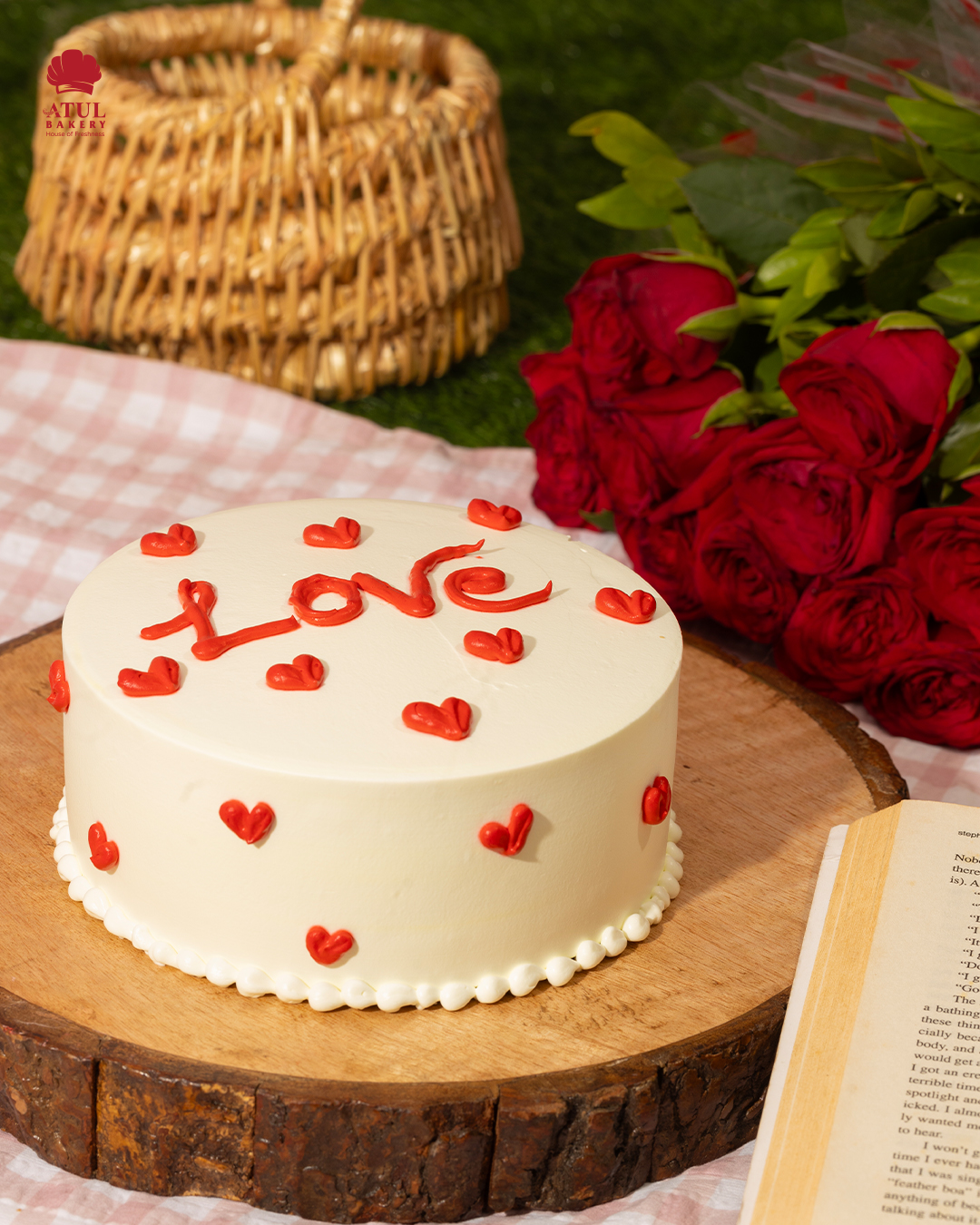 A white frosted round cake decorated with red hearts and the word “Love,” placed on a wooden base beside red roses, a picnic basket, and an open book — capturing a romantic picnic-themed setting by Atul Bakery.