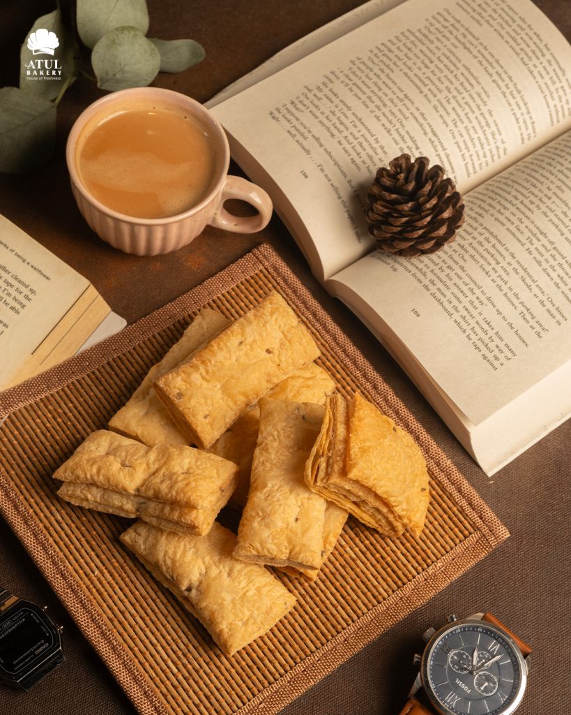 A cozy coffee setup with a cup of cappuccino, a plate of crispy jeera puffs from Atul Bakery, and open books — perfect for a relaxing snack break.