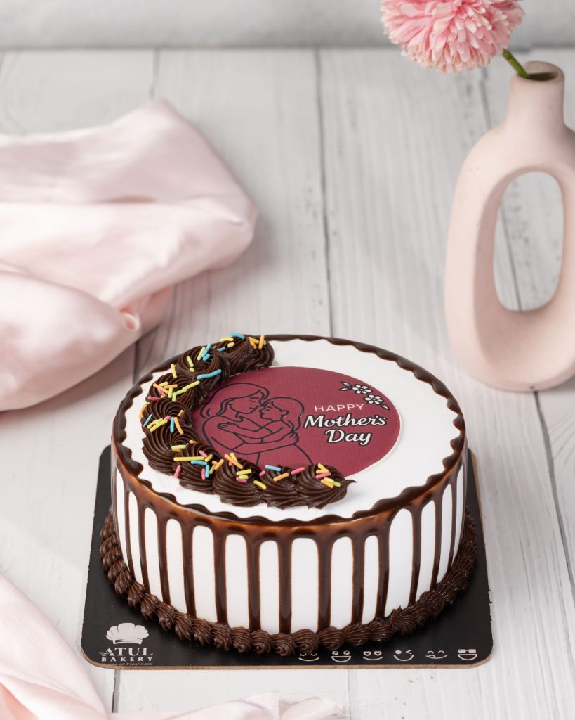 A beautifully decorated Mother’s Day cake from Atul Bakery with chocolate drips, frosting swirls, colorful sprinkles, and a pink edible topper featuring a mother holding a child.