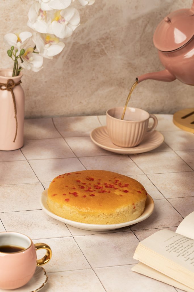A soft and spongy tutti frutti cake on a plate, surrounded by a pink teacup, teapot pouring tea, and an open book — creating a cozy tea-time setup with pastel tones.